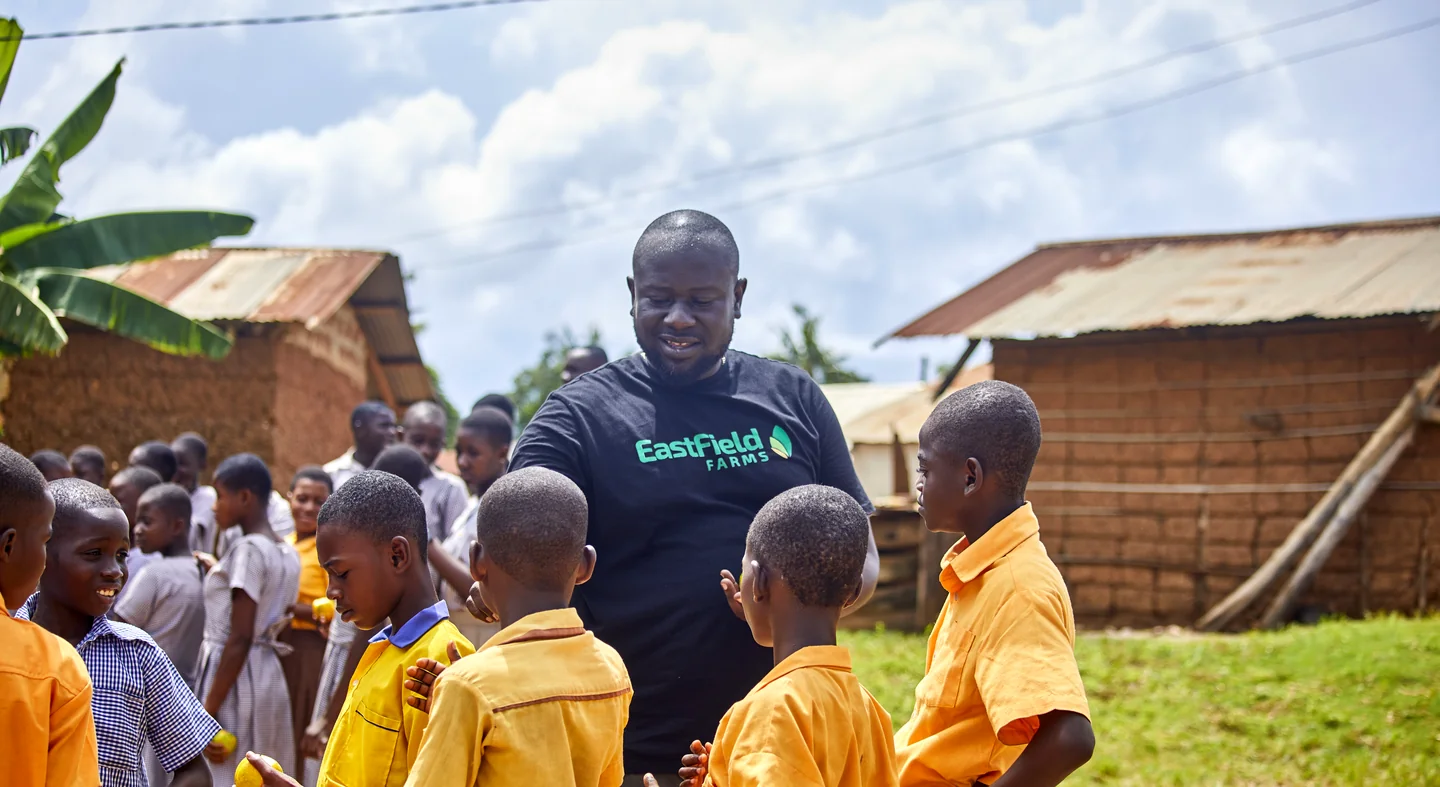 A visitor in an Eastfield Farms T-shirt talks with pupils in yellow uniforms outside their school.